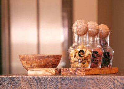 Exquisite Snacks im Spa- und Wellnessbereich: Frisch geröstete Nüsse im Glasdekanter mit Korkkugel auf Olivenholz, ideal für Gästeverwöhnung und luxuriöses Ambiente.