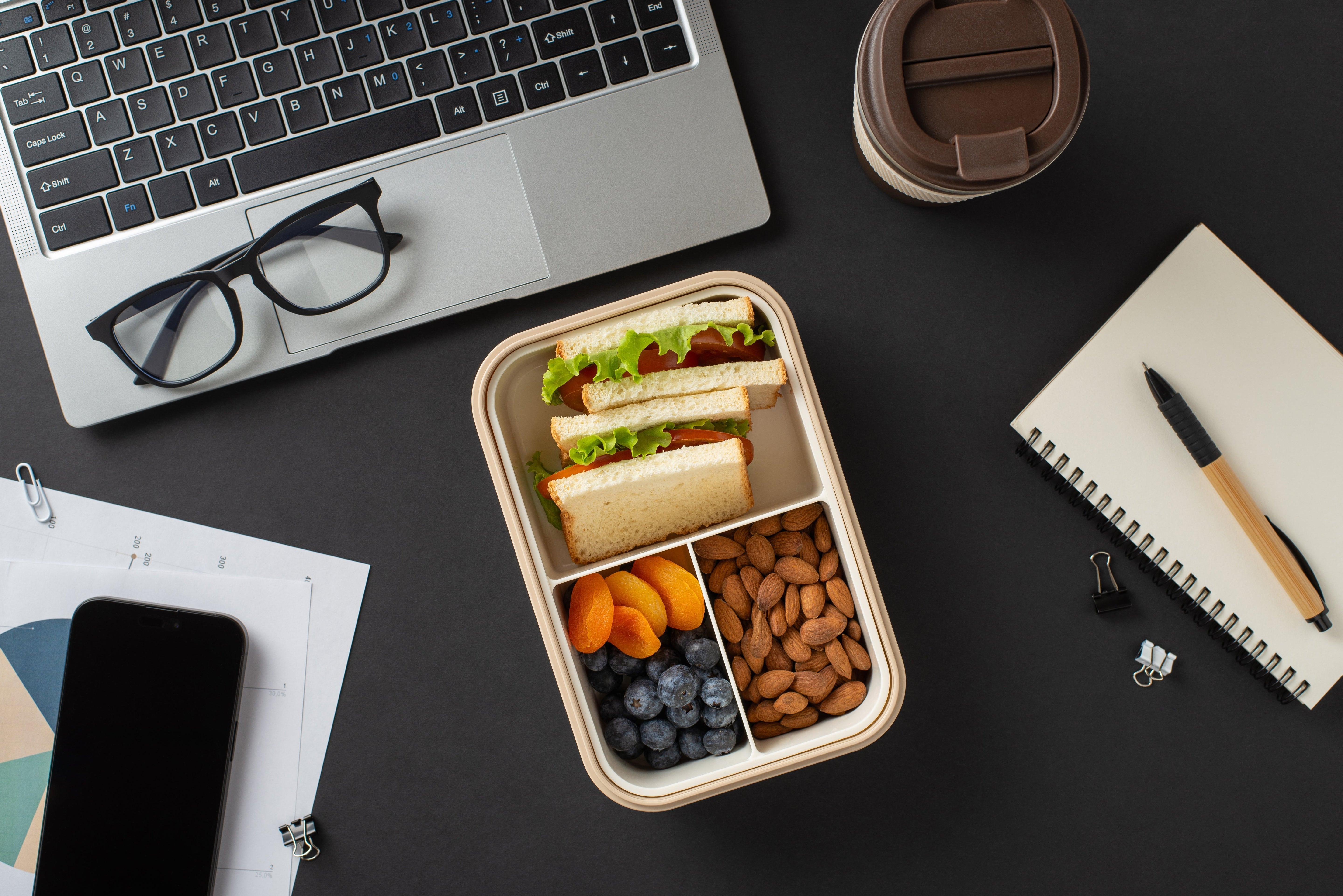 Snack-Box mit Nüssen, Obst, Sandwich auf einem Tisch mit Laptop, Block und Stift
