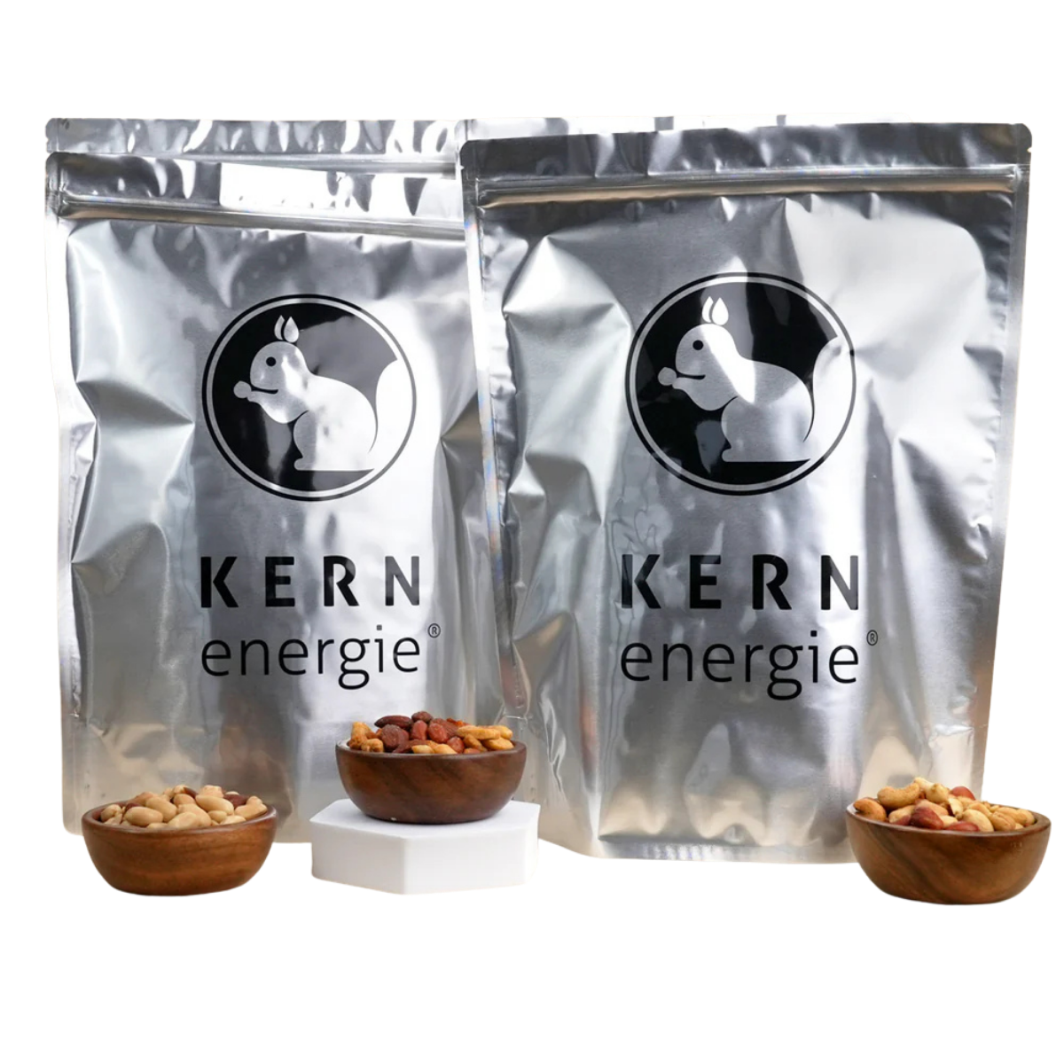 Zwei Alu KERNenergie Standbodenbeutel neben Schalen mit frisch gerösteten Nüssen.