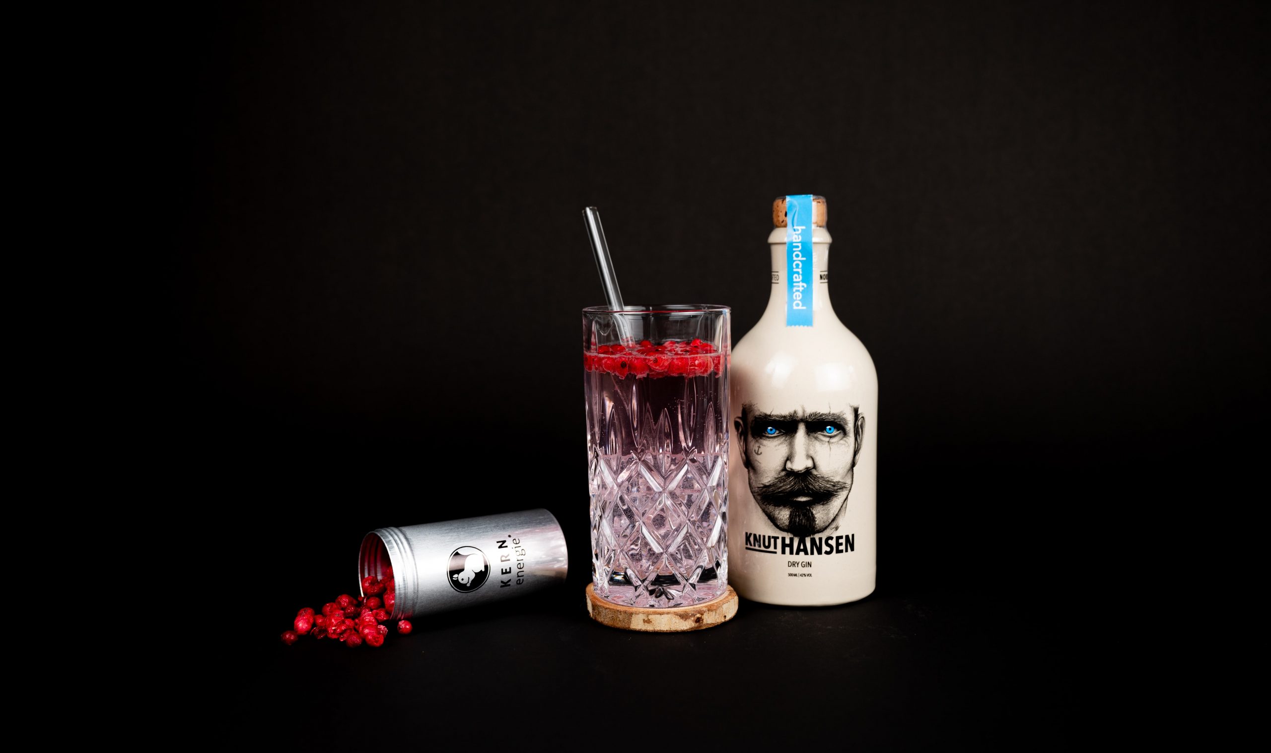 Gefülltes Gin Glas mit Strohhalm auf Holzuntersetzer zwischen einer Dose KERNenergie Gin Botanicals und einer Flasche Knut Hansen Gin vor schwarzem Hintergrund